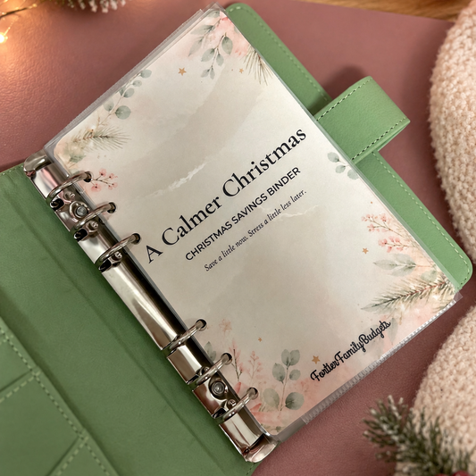 A green ring-bound binder with a clear cover displaying a printed insert that reads 'A Calmer Christmas Christmas Savings Binder' and 'Faithful Family Fundz,' styled on a pink surface with holiday lights, pine branches, and a candle in the background.