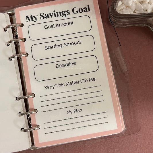 Savings goal planning sheet in a notebook with a glass container of rocks on a brown surface.