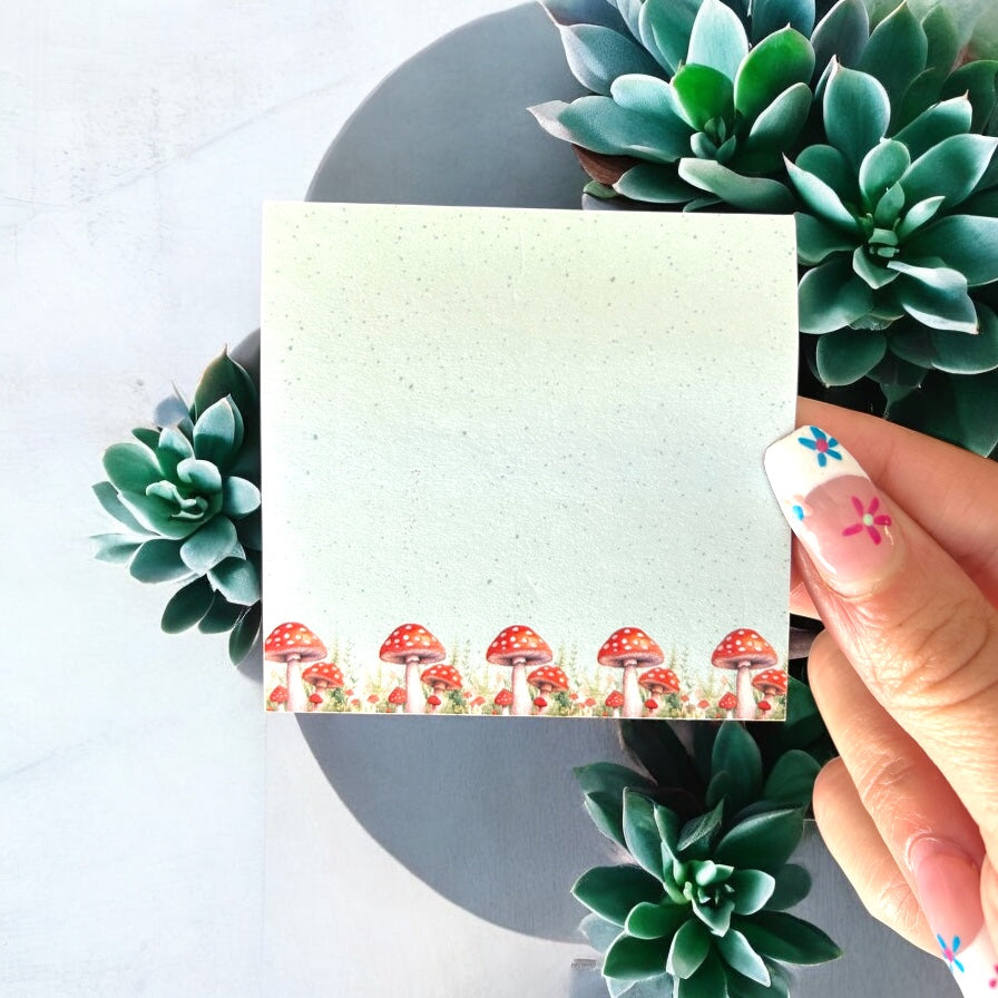 A hand holding a sticky note with a whimsical woodland mushroom design, with succulent plants in the background.