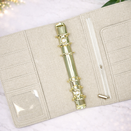 Beige fabric-bound notebook with gold rings on a white surface with green leaves.