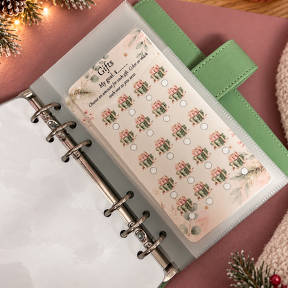 A close-up of a clear plastic binder page holding a decorative sticker sheet labeled 'Gifts' with repeated green gift boxes and small circular checkboxes, placed inside a green ring binder on a pink surface with holiday lights and pine decor visible.