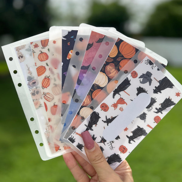 Hand holding a set of Halloween-themed cash envelopes with pumpkins and cats against a blurred outdoor background.