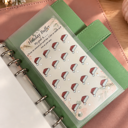 A green ring binder planner shown open on a pink surface, with a clear pocket holding a sheet of holiday-themed planner stickers featuring Santa hats. The scene is styled with warm lighting, a candle, and decorative evergreen branches in the background.