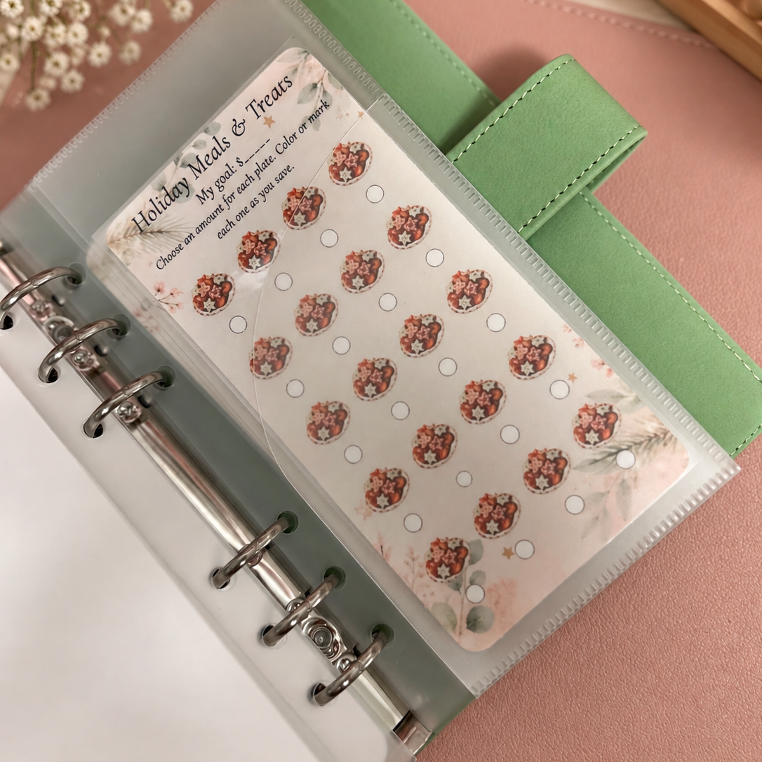 A clear plastic binder page holding a sheet of decorative holiday treats planner stickers, shown inside a green ring binder on a pink surface with small white flowers and a candle in the background.