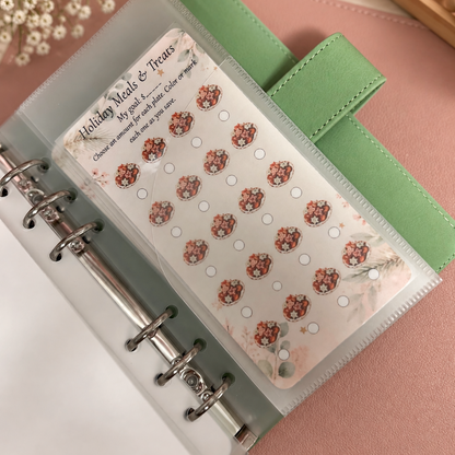 A clear plastic binder page holding a sheet of decorative holiday treats planner stickers, shown inside a green ring binder on a pink surface with small white flowers and a candle in the background.
