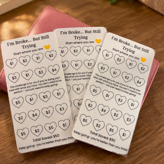 Three 'I'm Broke... But Still Trying' saving challenge cards on a wooden surface with a pink wallet.