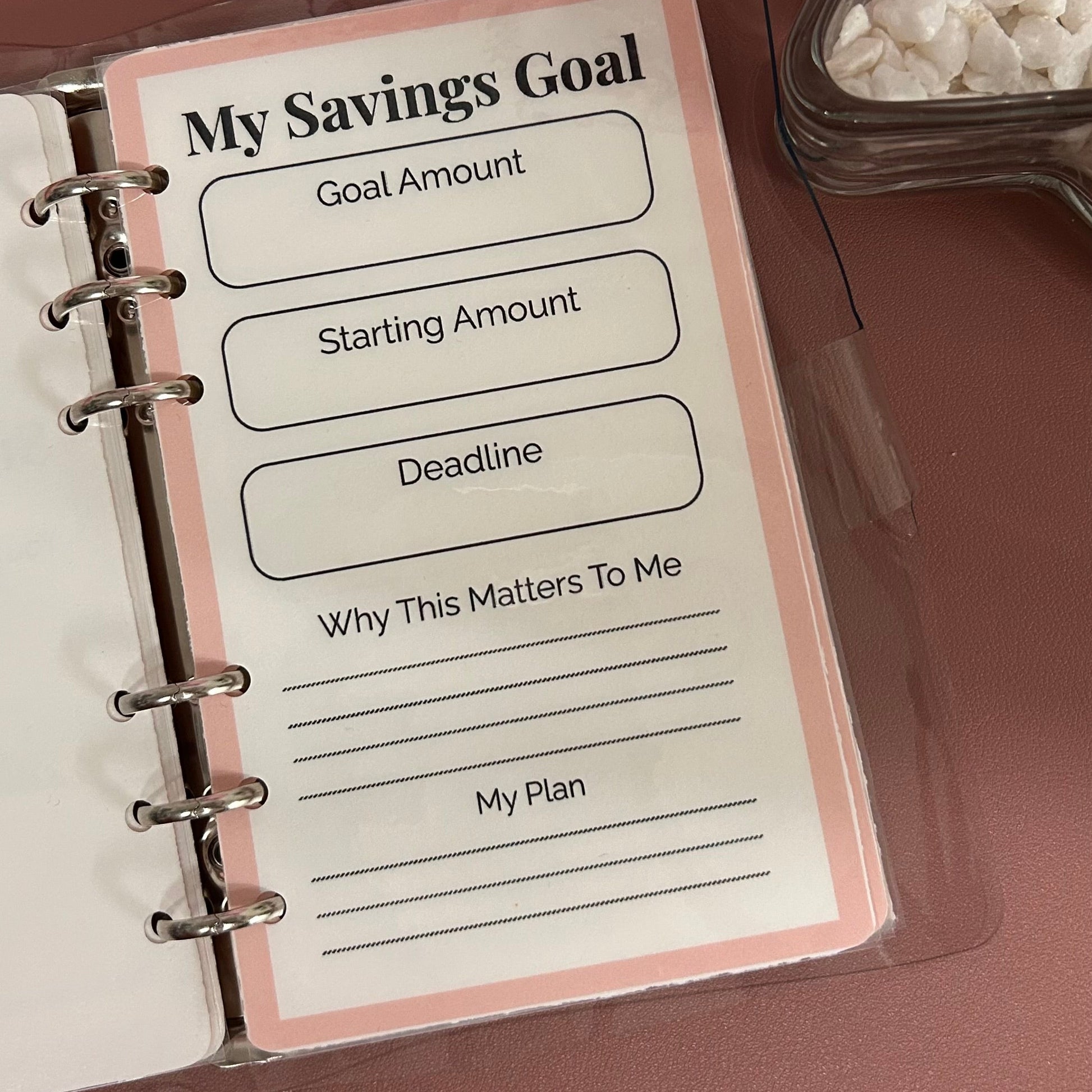 Savings goal planning sheet in a notebook with a glass container of rocks on a brown surface.