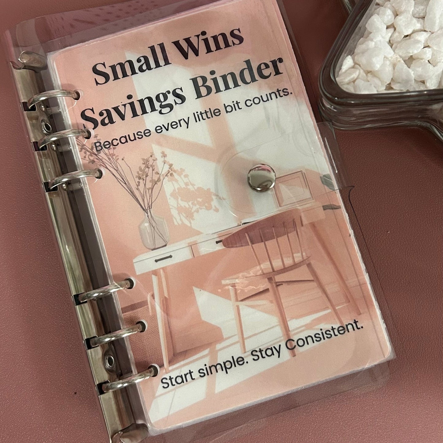 Small Wins Savings Binder on a wooden surface with a glass container of coins.