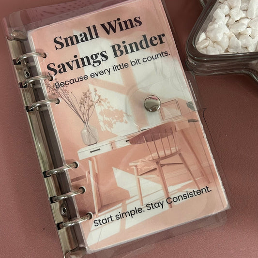 Small Wins Savings Binder on a wooden surface with a glass container of coins.