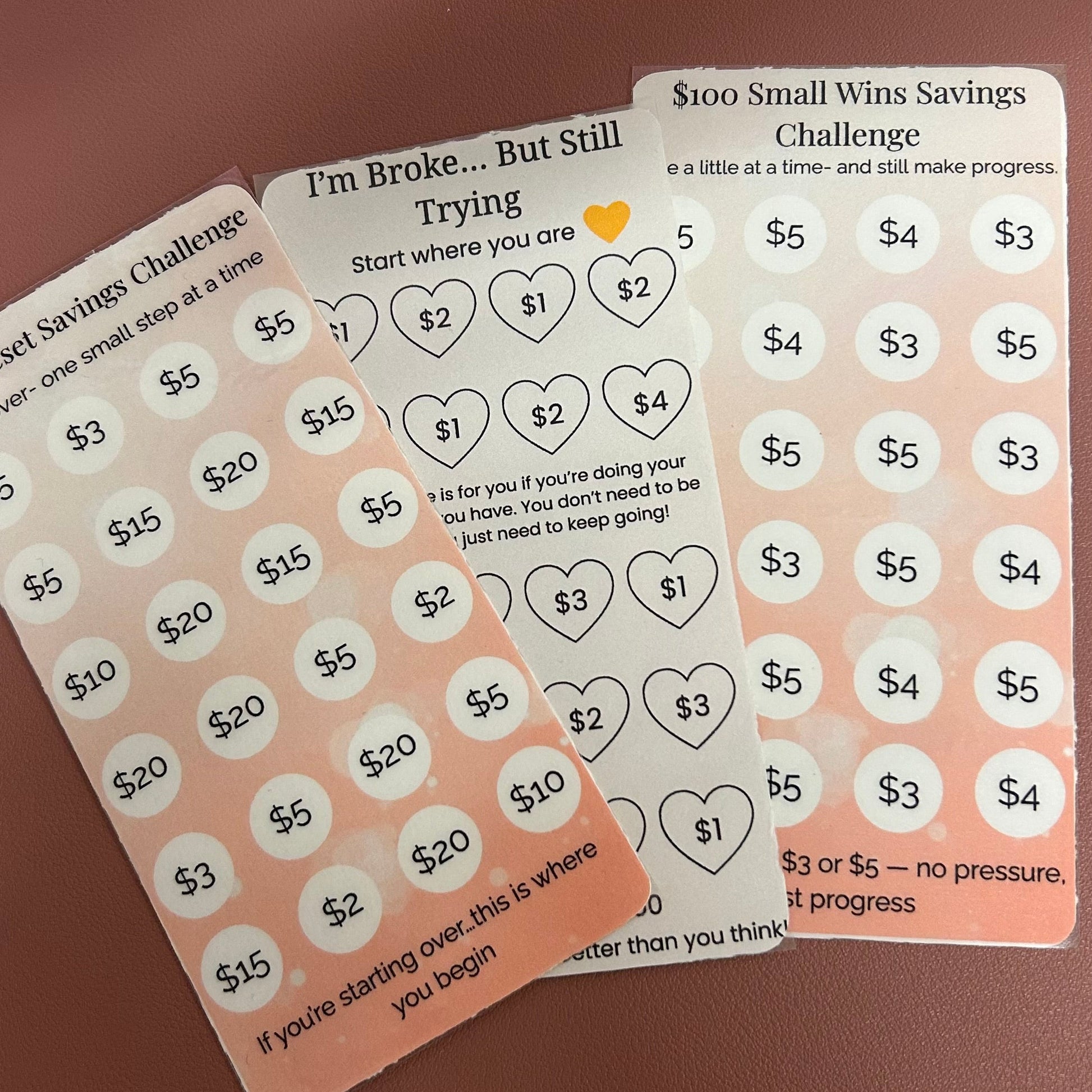 Three motivational saving challenge cards with heart and dollar amount stickers on a brown background.