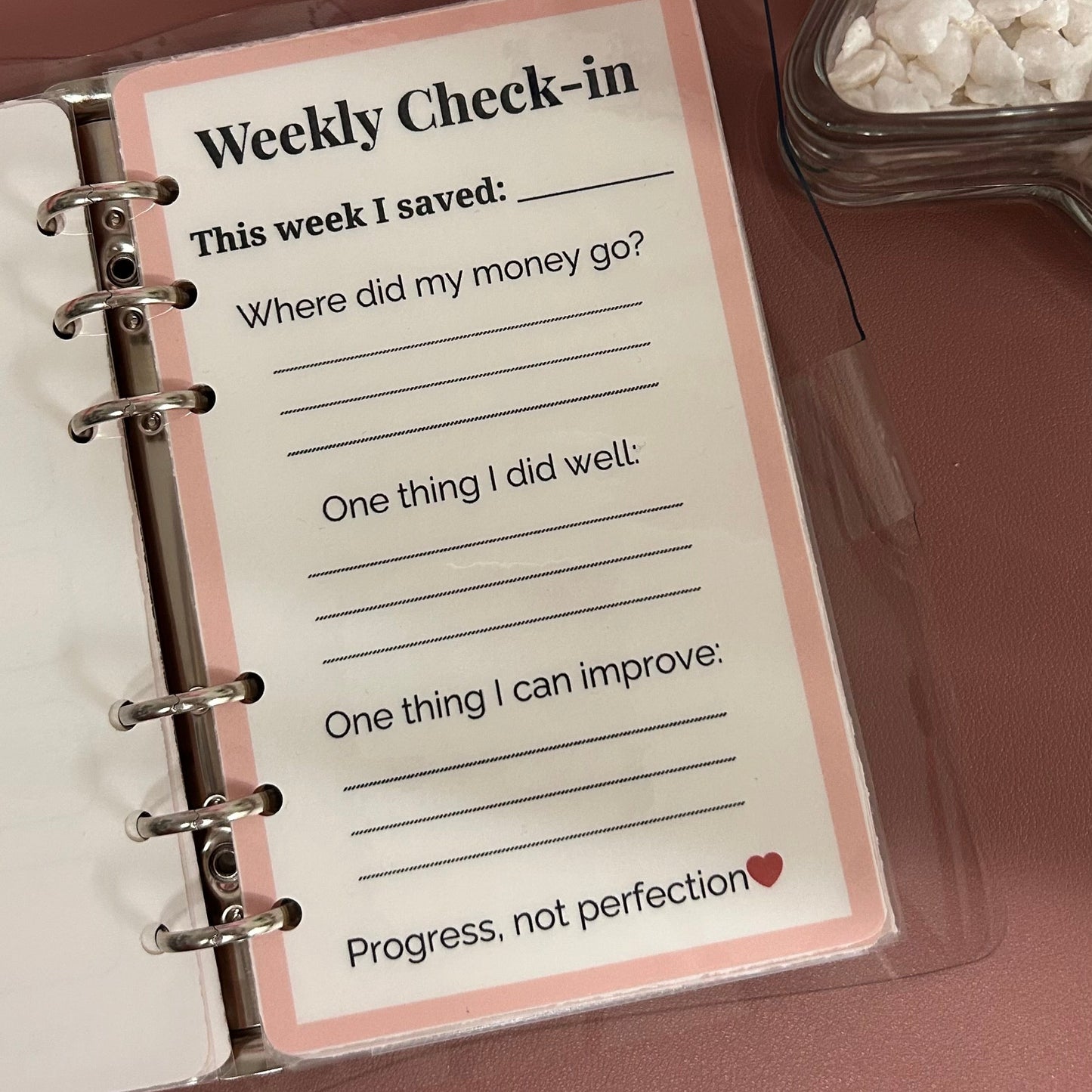 Weekly check-in card in a planner with a pink border on a brown surface.