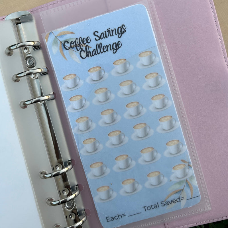 Pink planner with a sticker labeled 'Coffee Savings Challenge' on a wooden surface.