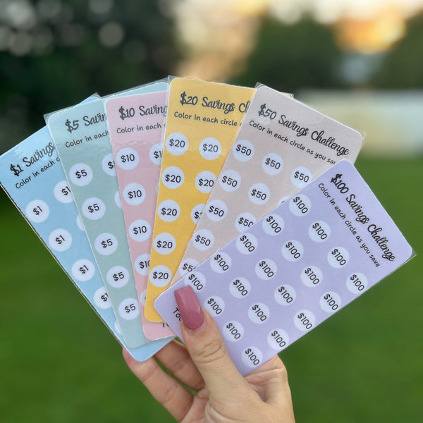 Hand holding five colorful saving challenge cards with numbers on a blurred natural background