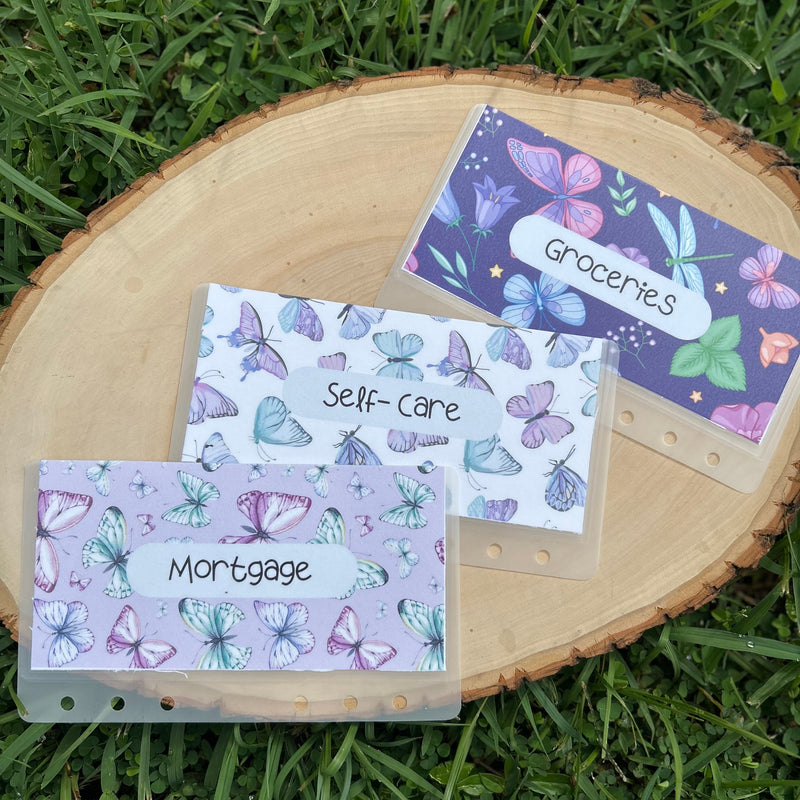 Set of three labeled envelopes with butterfly design on a wooden surface surrounded by grass.