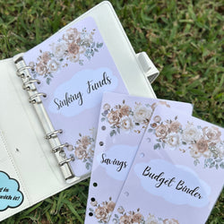 Floral-themed budget binder with 'Sinking Funds' and 'Savings' sections on a grass background.
