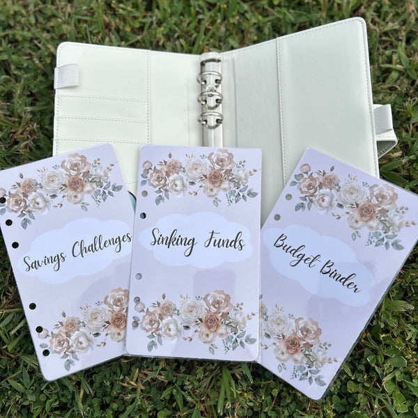 Three floral-themed budgeting sheets in front of an open planner on grass.