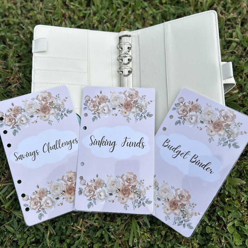 Three floral-themed budgeting sheets in front of an open planner on grass.