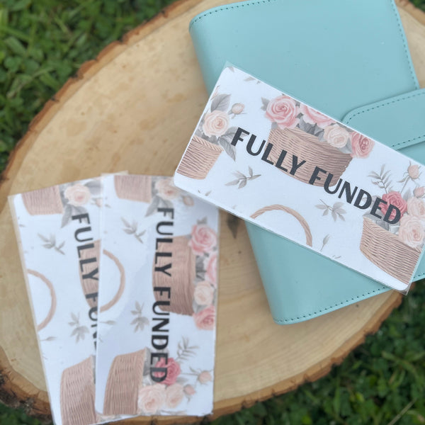 Two 'FULLY FUNDED' branded wallets on a wooden block with grass background