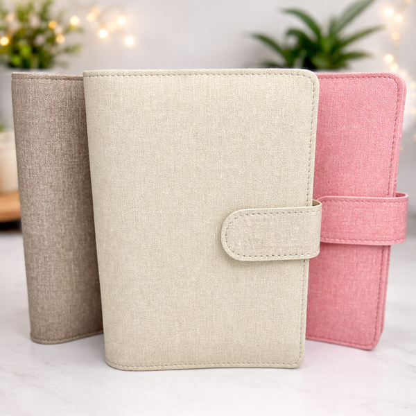 Three fabric-bound books in beige, pink, and another color on a white surface with a blurred background.
