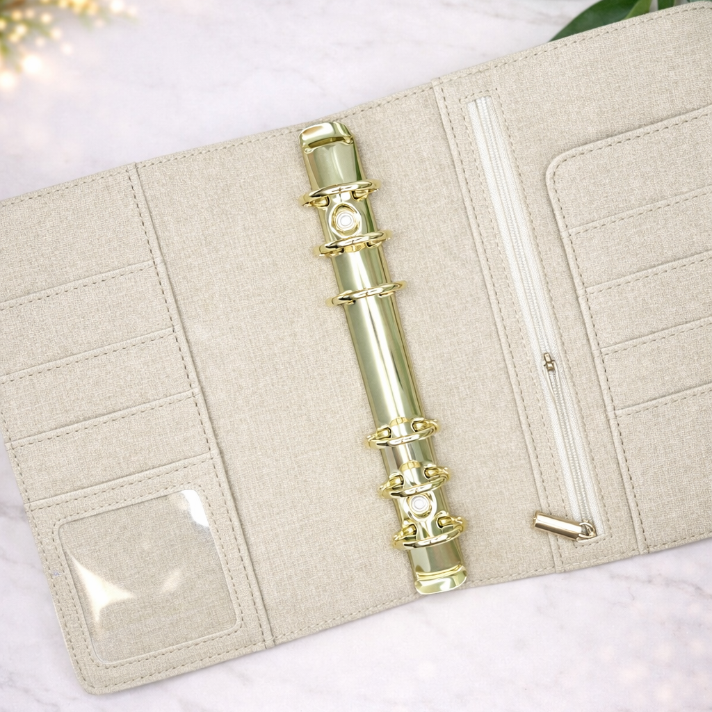 Beige fabric-bound notebook with gold rings on a white surface with green leaves.