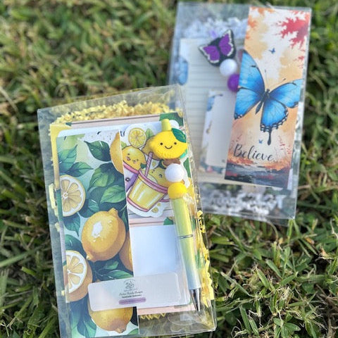Two decorative stationery items on grass, one with lemons and a yellow character, the other with a butterfly and text.